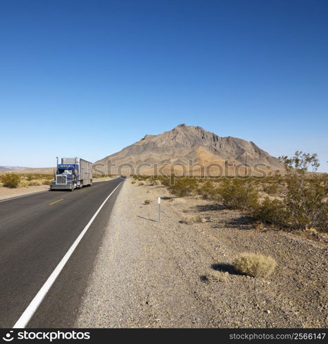 Tractor trailer driving on desert road with mountain in background.