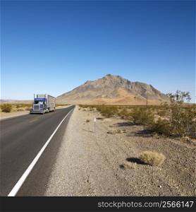 Tractor trailer driving on desert road with mountain in background.