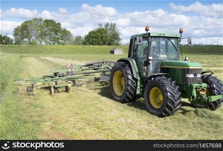 tractor with grass tedder during hay harvest in the netherlands under blue sky forms swaths in sunny field