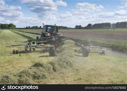 tractor with grass tedder during hay harvest in the netherlands under blue sky forms swaths in sunny field