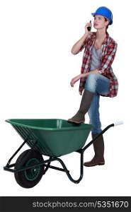 Tradeswoman speaking into a walkie-talkie and propping her foot up on a wheelbarrow