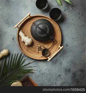 Traditional Asian tea ceremony arrangement. Iron teapot, cups, dried green tea leaves, ginger and tropical leaves over dark concrete background, flat lay, copy space. Traditional Asian tea ceremony arrangement, flat lay
