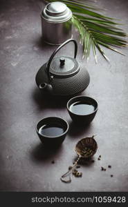 Traditional Asian tea ceremony arrangement. Iron teapot, cups, dried green tea leaves, ginger and tropical leaves over purple concrete background, top view, copy space. Traditional Asian tea ceremony arrangement, top view
