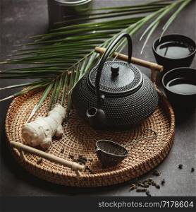 Traditional Asian tea ceremony arrangement. Iron teapot, cups, dried green tea leaves, ginger and tropical leaves over purple concrete background, top view, copy space. Traditional Asian tea ceremony arrangement, top view