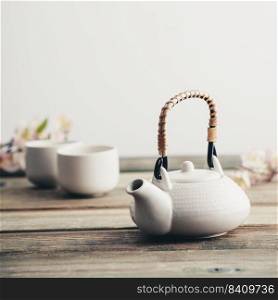 Traditional Asian tea ceremony arrangement. White teapot, cups, sakura flowers on wooden table against the white wall, selective focus, copy space