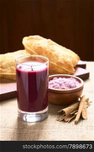 Traditional Bolivian Api, a purple corn beverage, with pastel (deep-fried pastry filled with cheese) in the back, photographed with natural light (Selective Focus, Focus on the front of the drink)