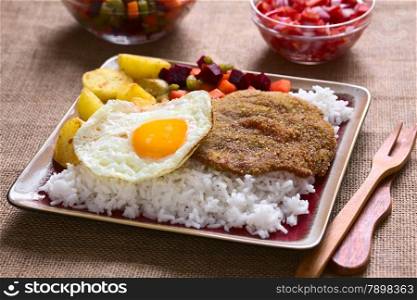 Traditional Bolivian dish called Silpancho, which is the name of the breaded flat, round piece of beef meat, served with fried egg, rice, fried potatoes, and vegetables (carrot, bean, beetroot), photographed with natural light (Selective Focus, Focus one third into the dish)