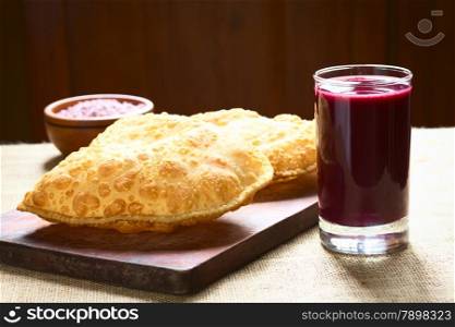 Traditional Bolivian snack called Pastel (deep-fried pastry filled with cheese) served with Api, a purple corn beverage on wooden board, photographed with natural light (Selective Focus, Focus on the front of the pastry and the glass)
