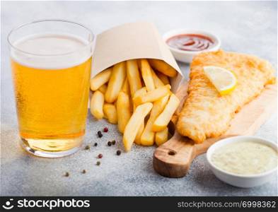 Traditional British Fish and Chips with tartar sauce abd glass of craft lager beer and tomato ketchup on chopping board on white stone background.
