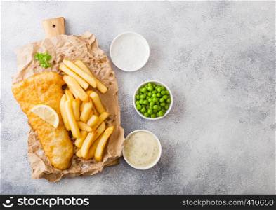 Traditional British Fish and Chips with tartar sauce on chopping board and green peas on white stone background. lemon slice and salt