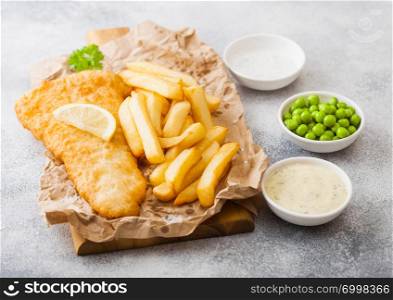 Traditional British Fish and Chips with tartar sauce on chopping board and green peas on white stone background. lemon slice and salt