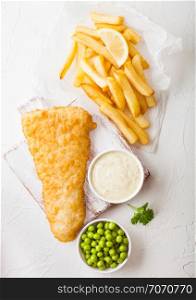 Traditional British Fish and Chips with tartar sauce on chopping board and green peas on white stone background.