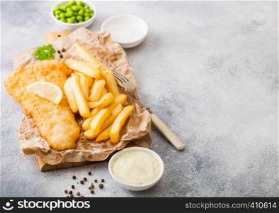 Traditional British Fish and Chips with tartar sauce on chopping board with fork and green peas on white stone background.