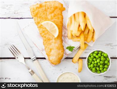 Traditional British Fish and Chips with tartar sauce on chopping board with fork and knife and green peas on white wood background.