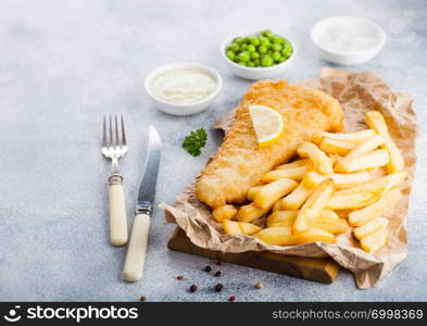 Traditional British Fish and Chips with tartar sauce on chopping board with fork and knife and green peas on white stone table background.
