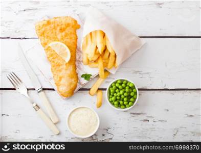 Traditional British Fish and Chips with tartar sauce on chopping board with fork and knife and green peas on white wood background.