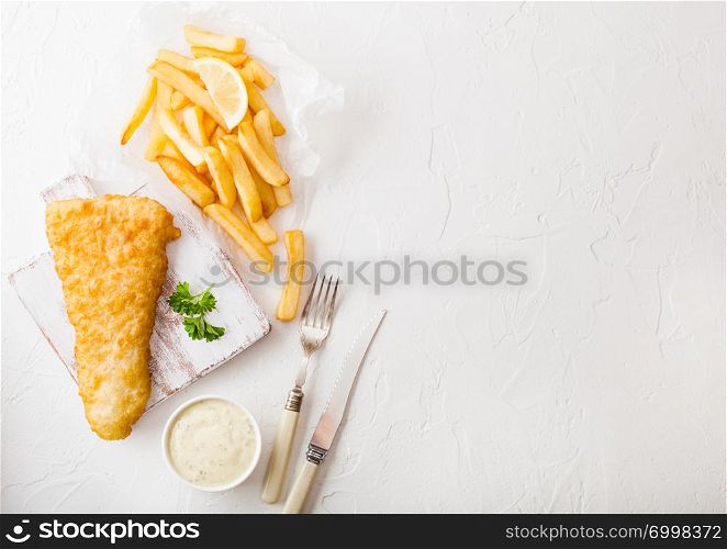 Traditional British Fish and Chips with tartar sauce on chopping board with fork and knife and slice of lemon on white stone table background.