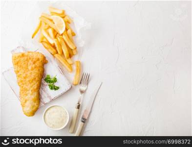 Traditional British Fish and Chips with tartar sauce on chopping board with fork and knife and slice of lemon on white stone table background.
