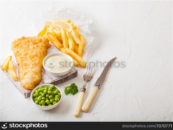 Traditional British Fish and Chips with tartar sauce on chopping board with fork and knife and green peas on white stone table background.