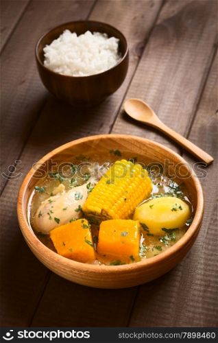 Traditional Chilean Cazuela de Pollo (or Cazuela de Ave) soup made of chicken, sweetcorn, pumpkin and potato, seasoned with fresh coriander served in wooden bowl with rice in the back (which is usually eaten with the soup), photographed on wood with natural light (Selective Focus, Focus one third into the soup)