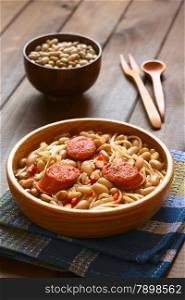 Traditional Chilean dish called Porotos con Riendas (English: beans with reins), made of cooked beans, linguine (flat spaghetti) and served with fried sausage, photographed on dark wood with natural light (Selective Focus, Focus on the two sausage pieces in the front)