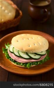 Traditional Chilean Hallulla bread roll prepared as a sandwich with lettuce, cold cut and cucumber served on wooden plate, photographed with natural light (Selective Focus, Focus on the front of the sandwich)