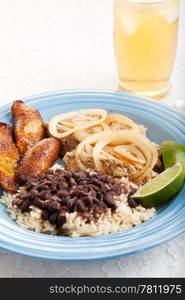 Traditional Cuban dinner of black beans and rice, roast pork, and fried plantains, with a glass of iced tea.