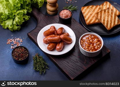 Traditional English breakfast with eggs, toast, sausages, beans, spices and herbs on a grey ceramic plate against a dark concrete background. Traditional English breakfast with eggs, toast, sausages, beans, spices and herbs on a grey ceramic plate