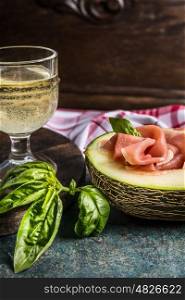 Traditional Italian lunch with prosciutto ham and melon, glass of wine and basilikum on dark rustic kitchen table, front view, close up
