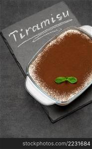 Traditional Italian Tiramisu dessert in glass baking dish on stone serving board with chalk inscription sign on concrete background or table. Traditional Italian Tiramisu dessert in glass baking dish on stone serving board with chalk inscription sign on concrete background