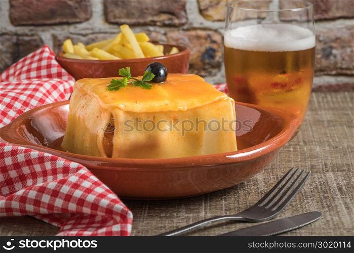 Traditional Portuguese snack food. Francesinha sandwich of bread, cheese, pork, ham, sausages, with tomato, beer, sauce and french fries. With a glass of beer and potatoes. On wooden table