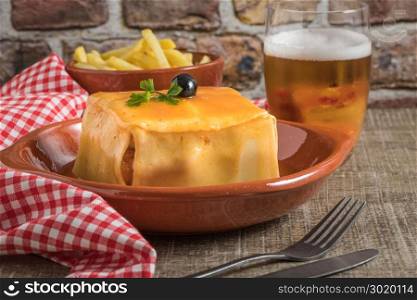 Traditional Portuguese snack food. Francesinha sandwich of bread, cheese, pork, ham, sausages, with tomato, beer, sauce and french fries. With a glass of beer and potatoes. On wooden table