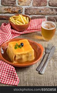 Traditional Portuguese snack food. Francesinha sandwich of bread cheese pork ham sausages with tomato beer sauce and french fries. With a glass of beer and potatoes. On wooden table