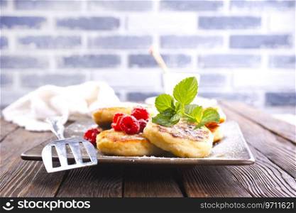 Traditional sweet syrniki or cottage cheese pancakes served with sweet cream and fresh raspberries in a white plate over table.