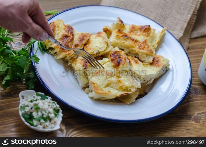 Traditional turkish borek in a enamel dish on wooden table.Woman's hand taking borek with fork.