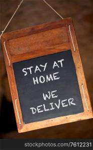 Traditional wooden blackboard or chalkboard sign hanging in a restaurant, cafe, store or shop saying Stay At Home We Deliver during Coronavirus COVID-19 Pandemic lockdown