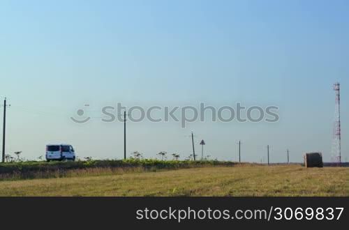 Traffic in the countryside. Cars going by the field with hay rolls against clear blue sky
