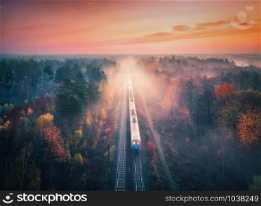 Train in beautiful forest in fog at sunrise in autumn. Aerial view of commuter train in fall. Colorful landscape with railroad, foggy trees, orange leaves, red sky and mist. Top view. Railway station