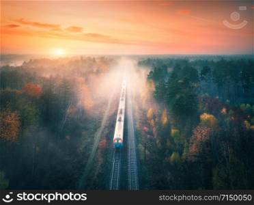 Train in beautiful forest in fog at sunset in autumn. Aerial view of commuter train in fall. Colorful landscape with railroad, foggy trees, orange leaves, red sky and mist. Top view. Railway station