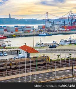 Train on railroad by comercial port with ships and containers, 25 de Abril Bridge over Tagus river at sunset, Christ the King monument on background, Lisbon, Portugal