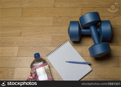 Training, exercise, cheerfulness and health - two plastic dumbbells, a notebook, mineral water and a pen on the wooden floor. The concept of a healthy lifestyle, fitness.. Training, exercise, cheerfulness and health - two plastic dumbbells, a notebook, mineral water and a pen on the wooden floor.
