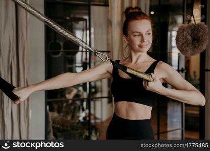 Training time of pilates female instructor on cadillac reformer, showing arm strengh exercises on machine with pretty smile during training in fitness studio. Healthy lifestyle and wellness concept. Training time of healthy happy ginger woman, exercising on cadillac reformer