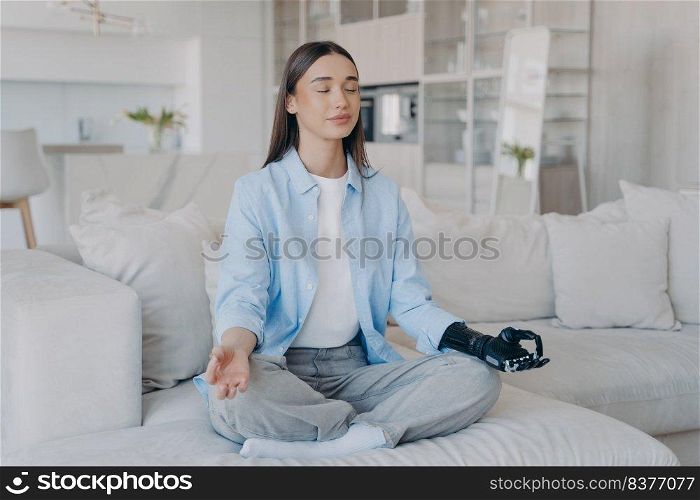 Tranquil disabled woman is sitting in lotus pose on couch with her eyes closed. Attractive caucasian girl is&utee. Lady has myoelectric cyber prosthesis. Modern artificial limb with sensor control.. Tranquil disabled woman is sitting in lotus pose. Modern artificial limb with sensor control.