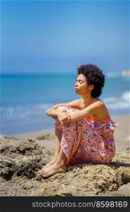 Tranquil ethnic female in maxi dress with curly hair and closed eyes, embracing knees while sitting on stones against waving sea and blue sky on summer weekend day on beach. Calm black woman resting against sea