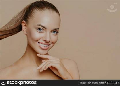 Tranquil undressed woman with healthy perfect skin, dark hair combed in pony tail, enjoys facial treatments, has well cared complexion, stands against beige studio wall, empty space. Skin care concept