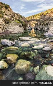 Translucent flowing river passing below ancient arched Genoese bridge at Asco in Corsica with colourful rocks, pebbles &amp; boulders in the foreground