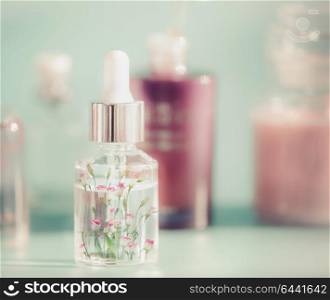 Transparent cosmetic bottle with liquid, pipette and little pink flowers. Floral essence or herbal extract serum on table, front view. Beauty and modern skin care concept