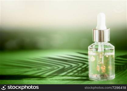 Transparent cosmetic bottle with pipette standing on green table with palm leaves shadow. Facial skin care concept. Natural vegan cosmetic. Serum or skin essential oil. Modern beauty trend.