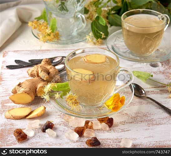 transparent cup of herbal linden tea and pieces of ginger on a white wooden board, next to pieces of sugar and twigs with green leaves