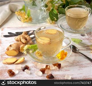 transparent cup of herbal linden tea and pieces of ginger on a white wooden board, next to pieces of sugar and twigs with green leaves